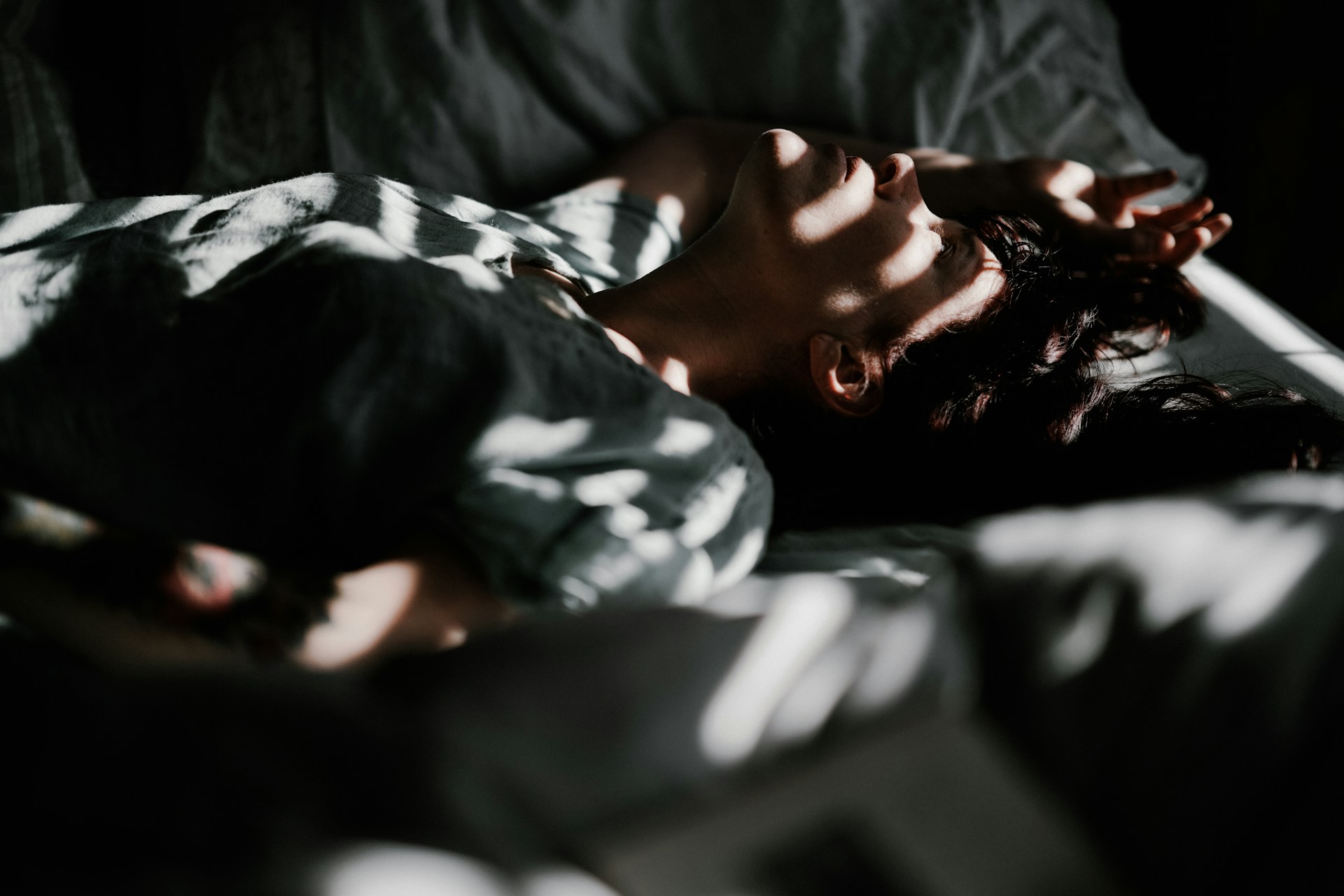 Frau liegt wach im Bett, gestreiftes Licht fällt auf ihr Gesicht, ihr Blick ist nachdenklich nach oben gerichtet