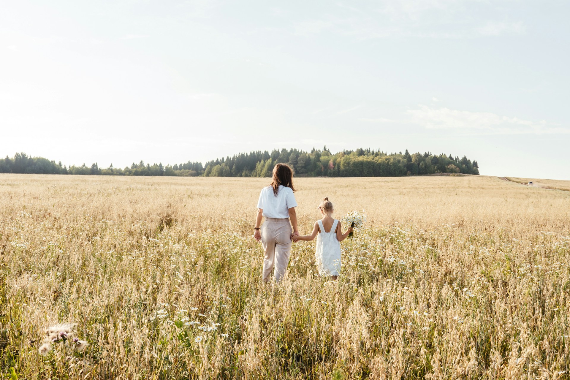 Mutter und Tochter gehen Hand in Hand durch ein sommerliches Wiesenfeld, die Tochter hält einen Blumenstrauß