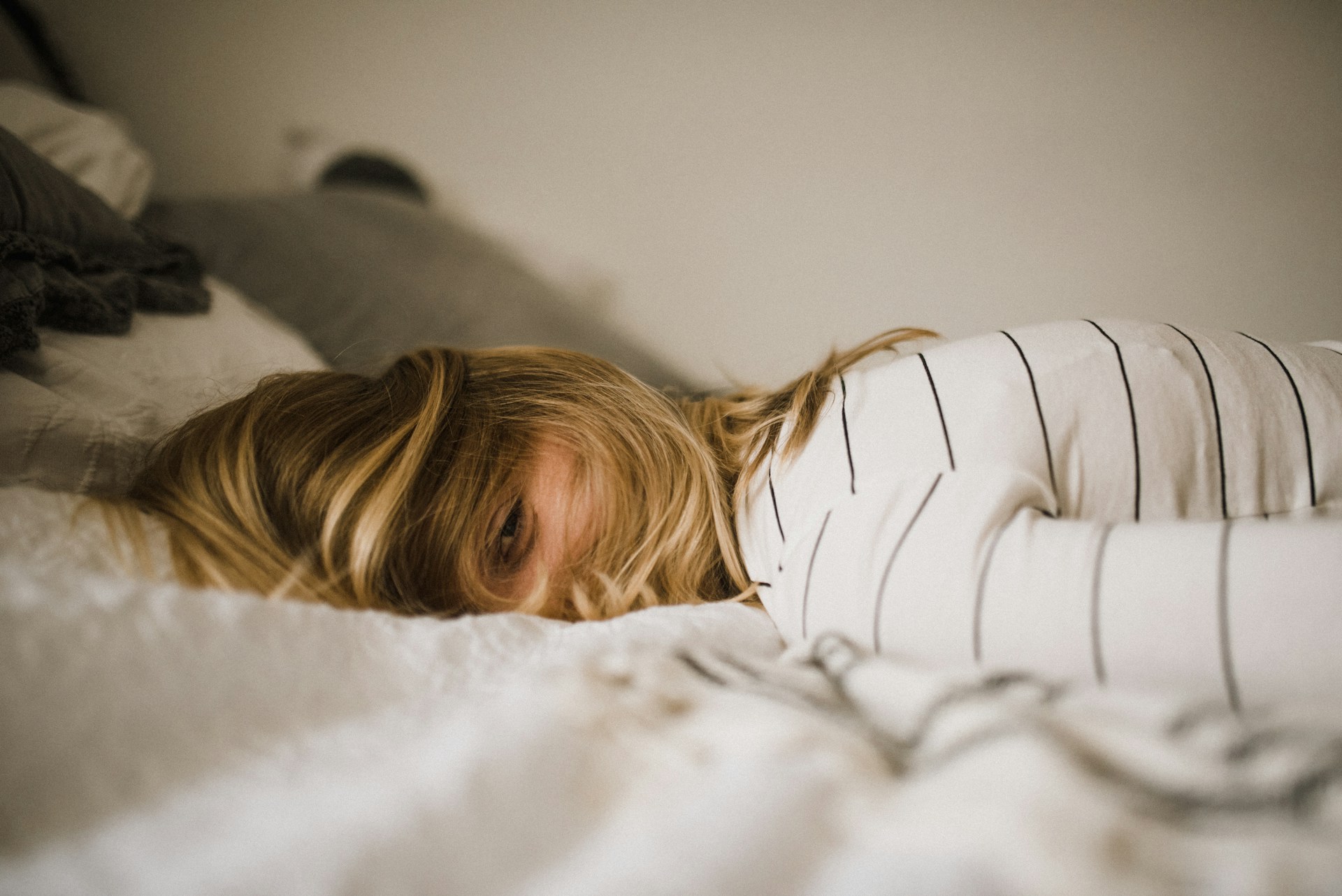 Frau liegt im Bett, ein müder Blick schaut durch die zerzausten Haare hindurch