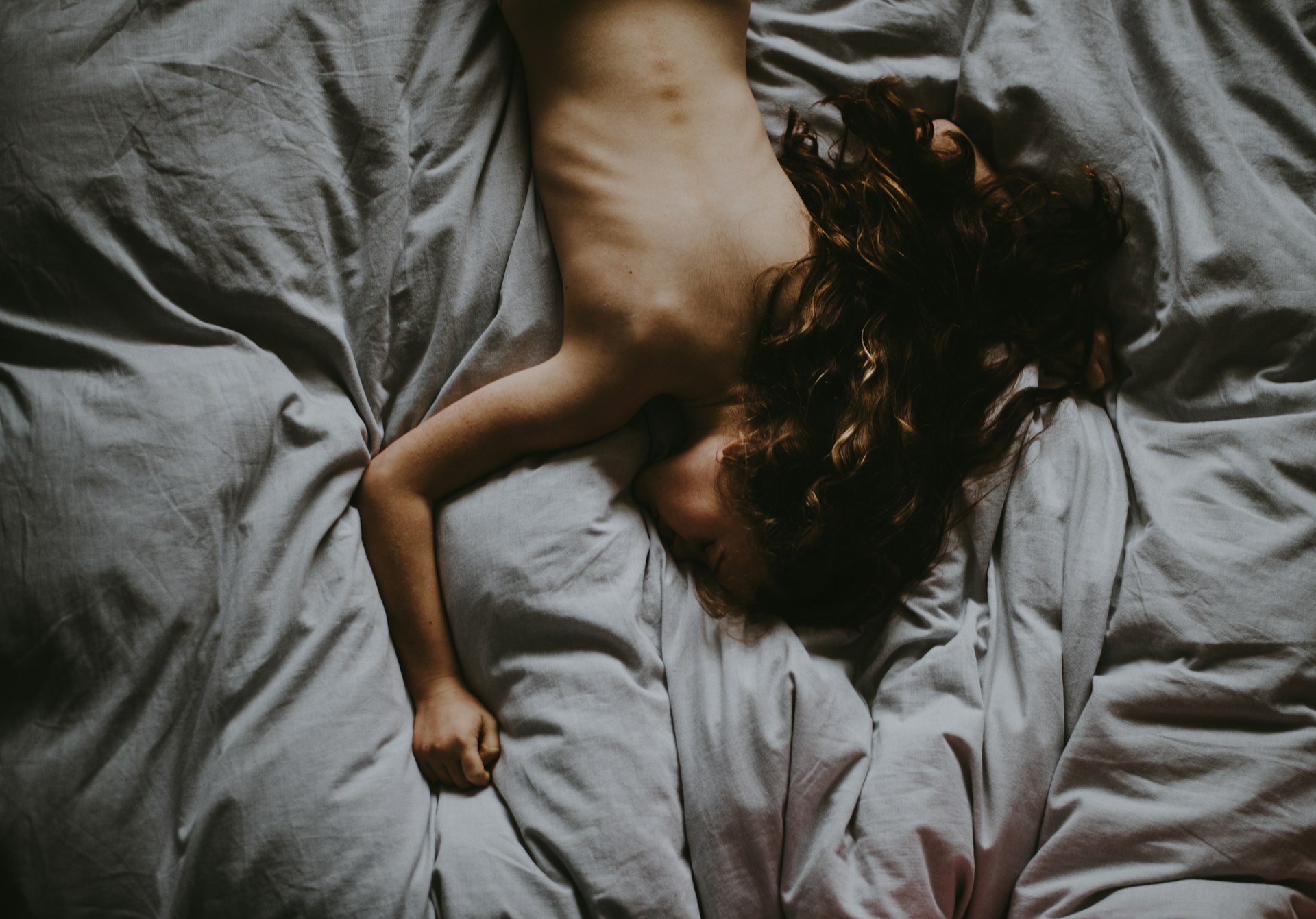 Frau liegt von oben fotografiert im Bett, Rücken und lockiges Haar sichtbar, gedämpftes Licht