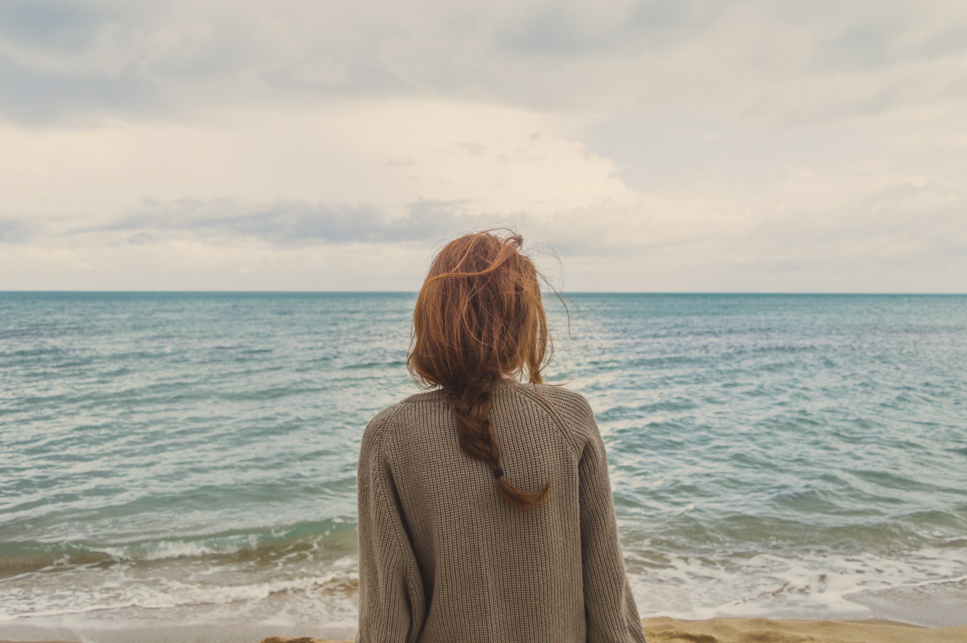 Frau von hinten mit geflochtenem Zopf blickt auf das Meer unter bewölktem Himmel