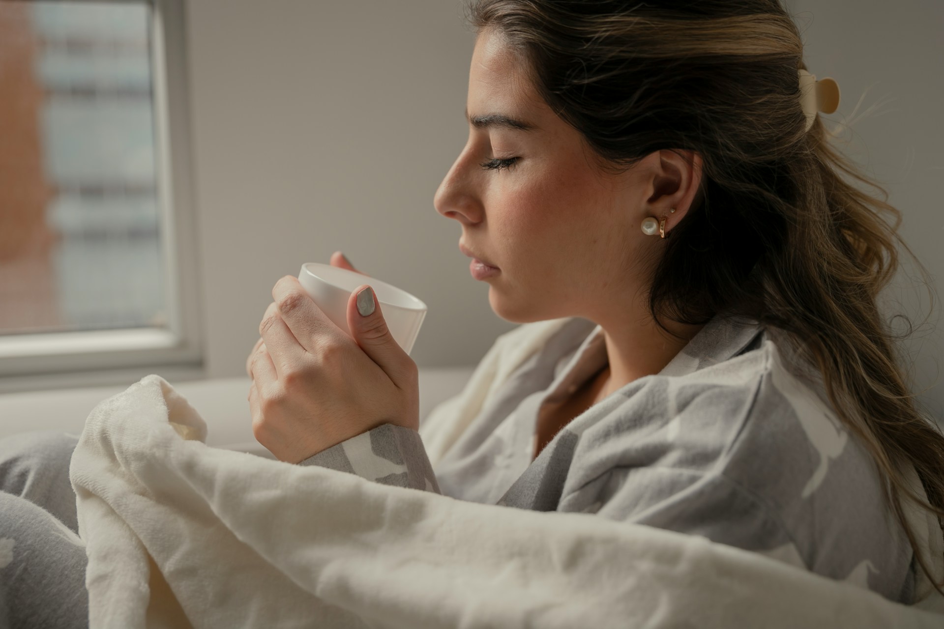 Frau in eine Decke gewickelt hält eine Tasse nah ans Gesicht, Augen geschlossen, weiches Fensterlicht