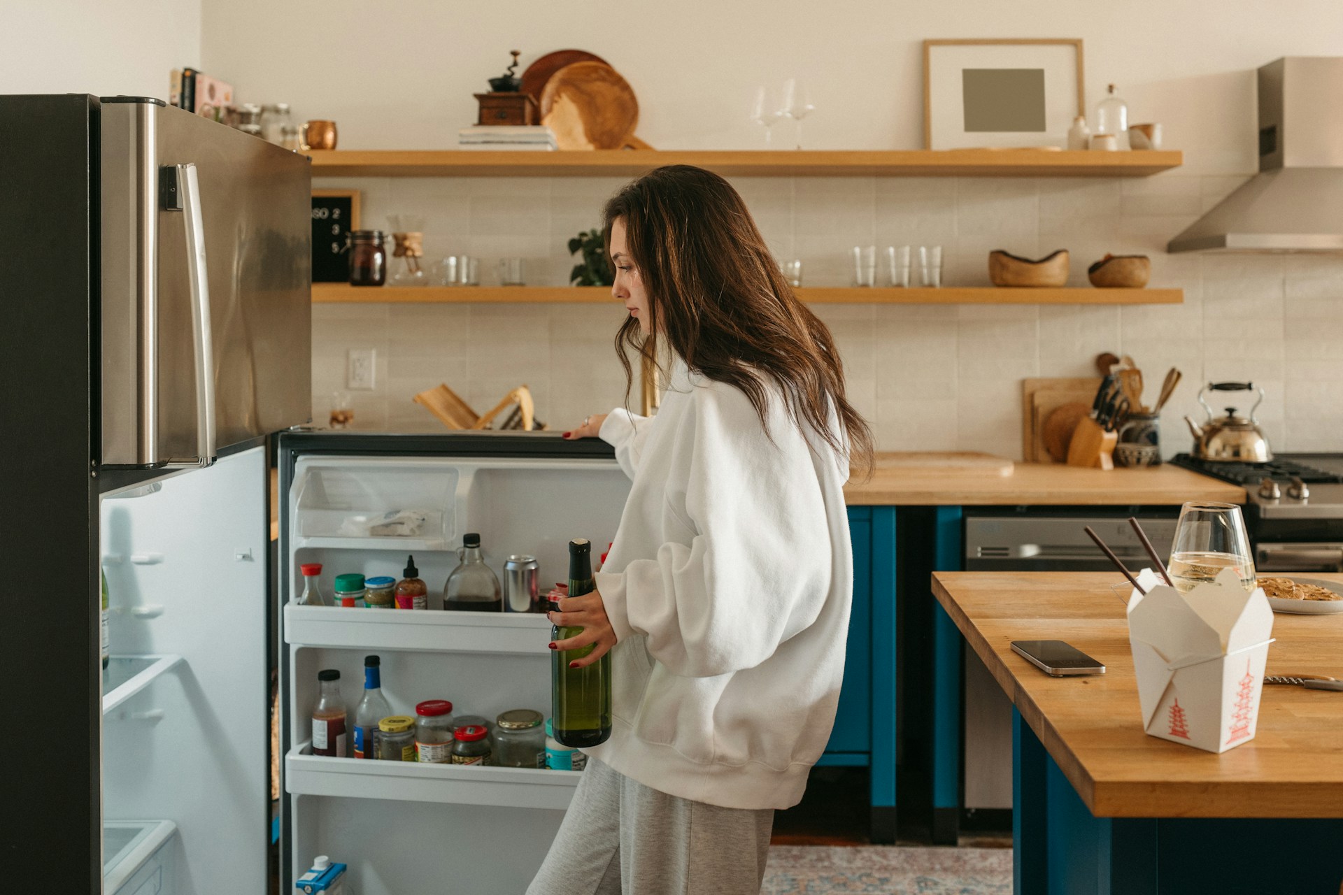 Frau steht nachdenklich vor dem offenen Kühlschrank und hält eine Flasche in der Hand
