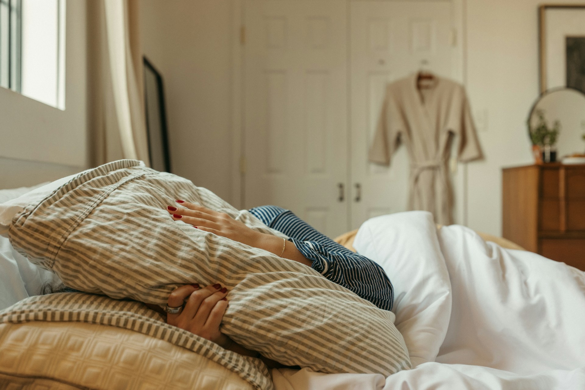 Frau liegt erschöpft im Bett, Gesicht unter der Bettdecke verborgen, nur Hände sichtbar, warmes Licht
